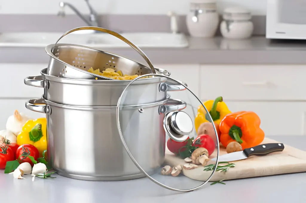 A set of stainless steel cookware on a kitchen counter, including pots with lids and a colander, surrounded by fresh vegetables and herbs.