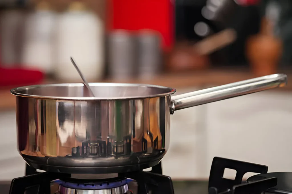 A gleaming stainless steel cooking pot with a long handle and a ladle inside it, placed on a gas stove burner.